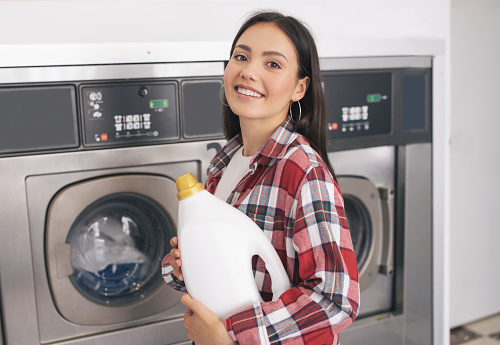 Laundrette Service. Cheerful Young Lady Holding Detergent Bottle Smiling To Camera, Advertising Laundromat Offer, Standing And Posing Near Industrial Washing Machines Indoors