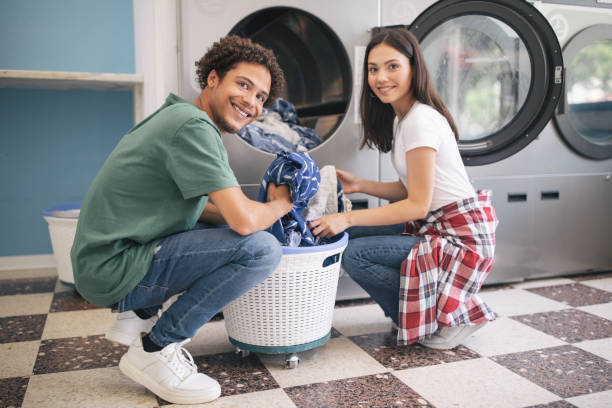 Happy Couple Doing Laundry Loading Washer Machine At Laundromat Room Happy Young Multicultural Couple Doing Laundry Together Loading Washer Machine At Laundromat Room. Guy Washing His Clothes And Posing Smiling At Camera. Public Laundrette Service laundry husband housework men stock pictures, royalty-free photos & images