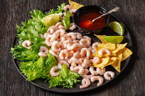 steamed shrimps with lettuce, lime wedges, corn chips and sweet chili sauce on black plate on dark wooden table, close-up