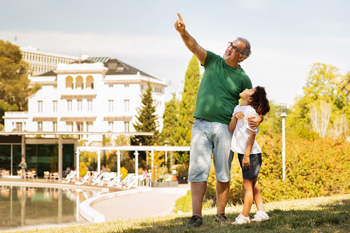 Smiling old caucasian grandfather hug little mixed race boy pointing finger at empty space, enjoy spare time, have fun outdoor. Rest, weekend and vacation in park together, family relationship