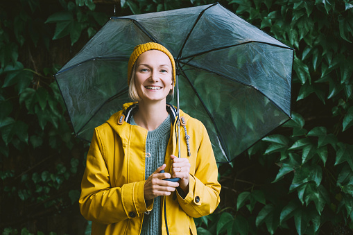 Happy woman wearing yellow raincoat holding transparent umbrella green leaves wall. Thoughtful young girl smiling outdoors at autumn season.