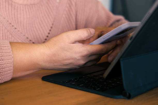 istock Female using phone looking at laptop or digital tablet device. Unrecognizable woman sitting at table in a cafe and use online banking with smartphone. Easy pay. App for sale in shop. Hands close up 1772433177