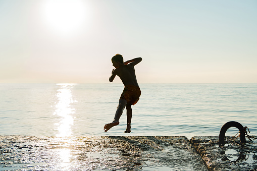 Silhouette of boy running along pier to jump into ocean water in early morning at sunrise. Happy child plays on beach at sea resort on summer vacation