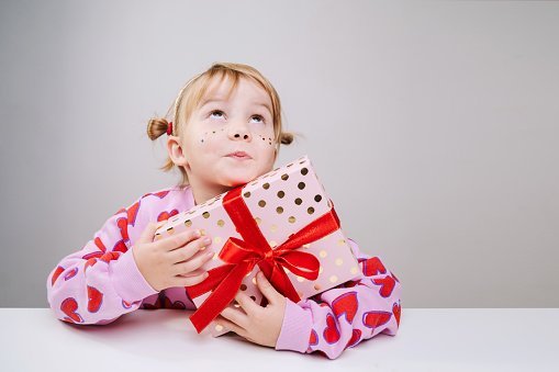 Cheerful little girl in pink sweatshirt with red hearts pattern holding gift box with red ribbon isolated over gray white background. Happy smiling small girl with surprise face posing in studio.