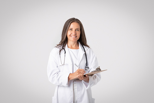 Comprehensive care planning. Cheerful senior woman doctor in white coat with stethoscope writing at clipboard on light background. Medical service, health care, prescription