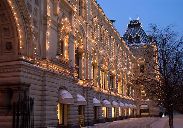 Budapest, Hungary - May, 2025A grand historic building with intricate architectural details, including multiple arches, towers, and ornate facades, stands illuminated by warm lights against the night sky. The multi-story structure features large windows and decorative elements, casting a glow on the surrounding area. In the foreground, a wet pavement reflects the lights, indicating recent rain, with a few indistinct pedestrians walking along the street.