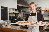 Welcoming female baker holding freshly baked almond croissants in  background of bakery shop.