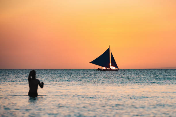 (messa a fuoco selettiva) splendida vista di una barca che naviga durante un bellissimo tramonto sullo sfondo e la silhouette di una persona sfocata che nuota in primo piano. spiaggia bianca, boracay, filippine. - nautical vessel philippines mindanao palawan foto e immagini stock