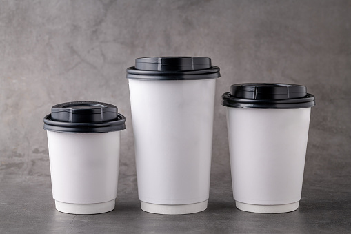 Three different sizes of white takeaway paper cups with black plastic lids isolated on gray background.