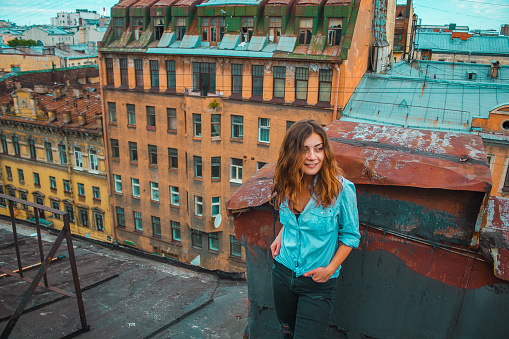 A smiling girl with brown hair stands on a roof overlooking an old building and historical street in Saint Petersburg, Russia. Traveler on an excursion. Horizontal cityscape with one woman