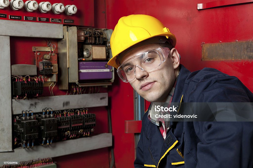 Patch panel inspector in hard hat facing camera by machinery Young engineer inspecting the main patch panel in his factory. Adult Stock Photo