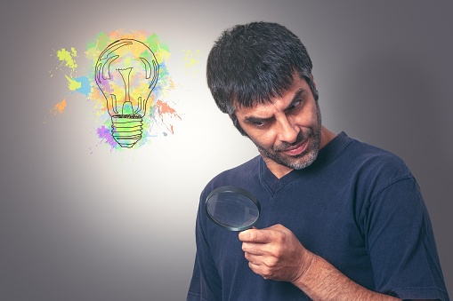 A mature man stands against a plain white backdrop while examining a magnifying glass in his hands