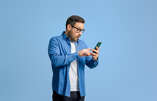 Shocked young businessman in eyeglasses reading bad news over mobile phone against blue background