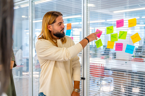 Creative young man next to a glass with adhesive notes explaining ideas to colleagues