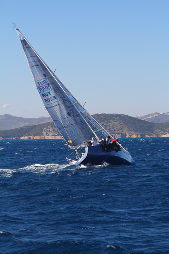 Bodrum,Mugla, Turkey. February 09, 2020:  sailor team driving sail boat in motion, sailboat wheeling with water splashes, mountains and seascape on background. Sailboats sail in windy weather in the blue waters of the Aegean Sea, on the shores of the famous holiday destination Bodrum