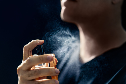 Fragrance spray. Man and perfume. Applying cologne. Scent water. Skin care, beauty product and male cosmetics concept. Holding elegant smell bottle in hand. Dark blue background. Young gentleman.
