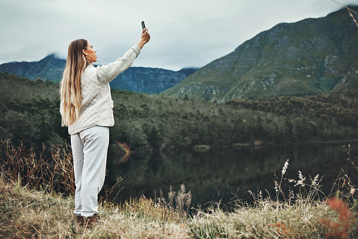 Woman, selfie and forest, mountains or nature in search of network, connection or internet outdoor. Young person camping, travel or hiking with adventure photography or mobile for location by lake