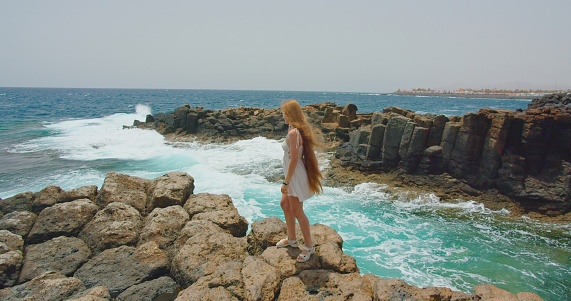 Fuerteventura, Canary Islands. Attractive multiracial woman enjoying paradise, sunny day on clear azure ocean rocky seaside relaxing, wind blowing long hair. Sea waves crash foaming on volcanic cliffs. Slow motion. Adult tourist.