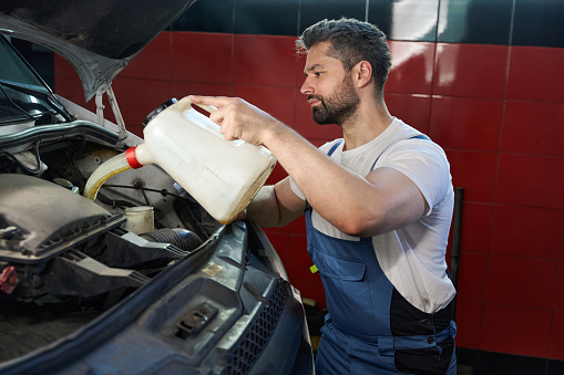 Concentrated automotive technician pouring motor oil from plastic tank into car engine while standing in front of open vehicle hood