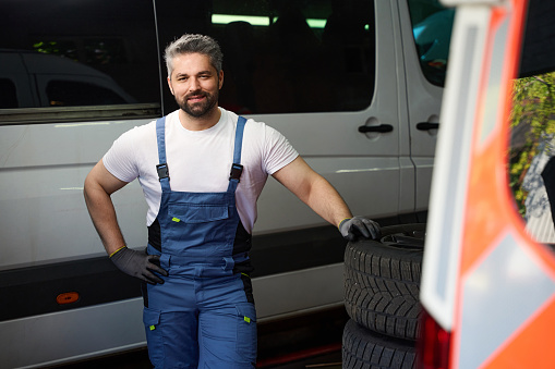 Pleased auto mechanic leaning on stack of car tyres while standing near van in garage