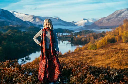 Woman wearing a Grant tartan traditional kilt and scarf above Loch Affric in the Highlands of Scotland. The distant hills are coated with the first snows of autumn.