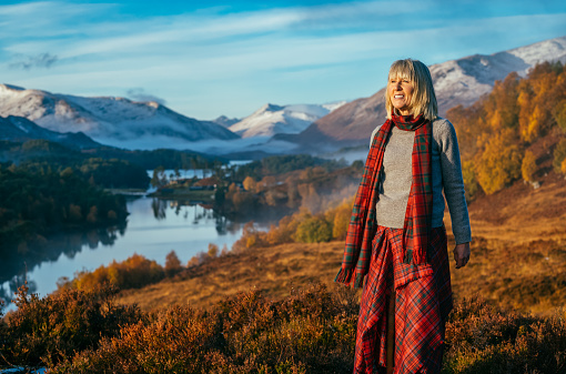 Woman wearing a Grant tartan traditional kilt and scarf above Loch Affric in the Highlands of Scotland. The distant hills are coated with the first snows of autumn.