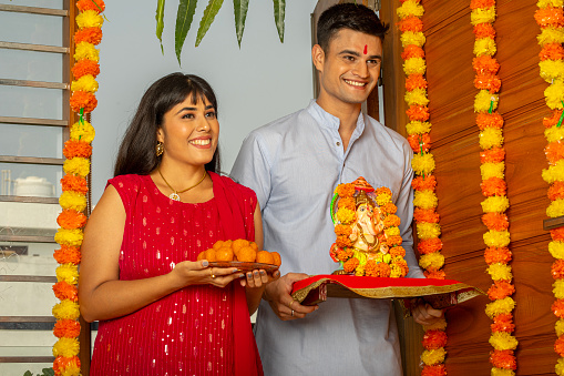 Happy young husband and wife with Ganesha statue and plate of laddoos standing at entrance of decorated house during religious festival
