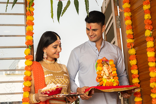 Happy young husband and wife with Ganesha statue and plate of flowers standing at entrance of decorated house during religious festival