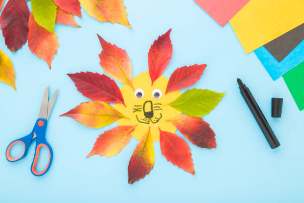 istock Smiling lion head shape created from colorful leaves and paper on light blue table background. Pastel color. Making autumn decorations. Closeup. Top down view. 1772745909