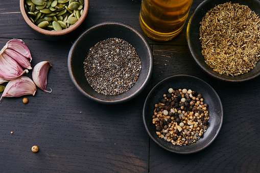 Dark mood variety of vegan herbs and spices, seasoning on a black wooden table top kitchen or restaurant table, pepper, coriander seeds, chia seeds, red pepper, garlic cloves, chili pepper, Indian nuts, cardamom, black sesame, salt, top view with a copy space