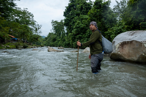 Man crosses a swift river using a wooden stick during a hike in the rainforest.
