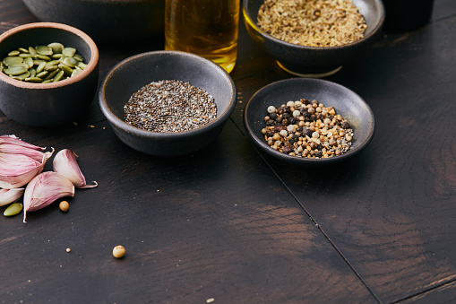 Dark mood variety of vegan herbs and spices, seasoning on a black wooden table top kitchen or restaurant table, pepper, coriander seeds, chia seeds, red pepper, garlic cloves, chili pepper, Indian nuts, cardamom, black sesame, salt, top view with a copy space