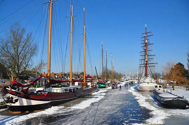 "winter scene, frozen river with ice skaters, old sailingships and blue sky, smaal harbor of Stavoren"