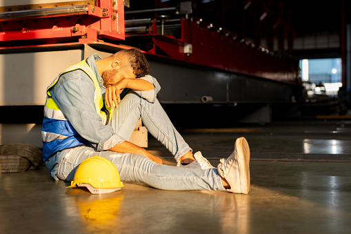 exhausted industrial worker has problem sitting on floor behind machine feeling sick headache injured pain, tired male technician foreman wiping sweat after stressed work in manufacturing factory