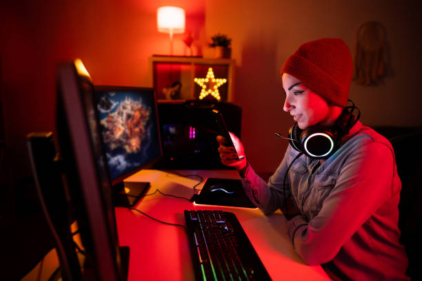 Young Caucasian female gamer and streamer, using mobile phone while playing video games on the computer in her LED-lit room