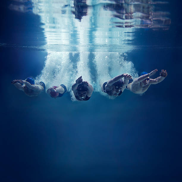 Five swimmers jumping together into water, underwater view Five, young swimmers are jumping in direction of the camera. This is underwater shoot. There are 3 girls and 2 boys. They wear caps, swimming goggles and swimwears. The swimmers just entered the water with hands in front of themselves. They are looking down. They leave behind them a lot of air bubbles. In the top of image you can see the surface of water and reflection of the scene. The background is clear, dark blue. There are no swimming pool elements. This is a square image. There is a lot of free space under the swimmers. coordination photos stock pictures, royalty-free photos & images