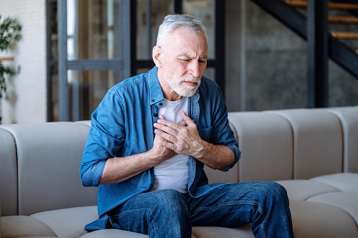 Asthma symptoms. Senior man having difficulties with breathing. Male sitting on sofa and holding hands on chest in living room at home. Chronic illness concept.