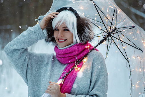 Woman, adorned in a white wig, thermal earmuffs, cozy sweater, and pink scarf. She holds a transparent umbrella embedded with twinkling lights, creating a magical winter atmosphere.