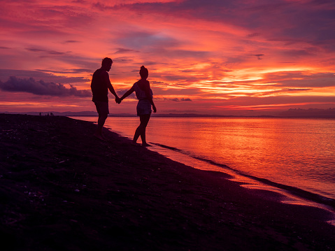 Classical Valentine Day scene with the silhouette of young couple holding hands while contemplating sunset at the beach in the island of Koh Phangan, Thailand. Lovers staring at sun, honeymoon concept