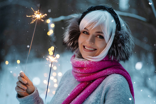 Woman, adorned in a white wig, thermal earmuffs, cozy sweater with sparklers during winter holidays.