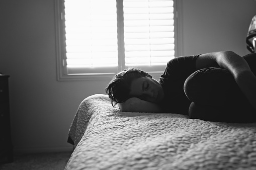 A woman lies on a bed, curled up around herself. Her eyes are closed.