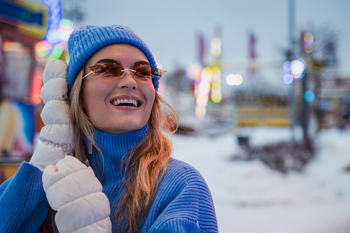 Cheerful and stylish woman, dressed in warm clothes and wearing sunglasses shaped like fire flames, is having fun in a snowy winter amusement park.