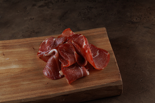 Italian prosciutto crudo or spanish jamon. Raw ham on wooden cutting board, on dark brown background. Ingredient High resolution. Copy space, side view