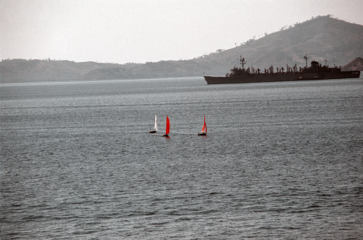 View of sailboats in Puget Sound with US Navy Sacramento-class fast combat support ship in silhouette in background (unidentified but likely USS Seattle (AOE-3)).