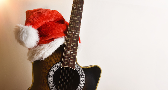 Acoustic guitar body with Santa hat leaning on an isolated white wall. Christmas guitar music concept.