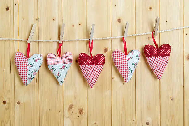 Five colored cotton hearts hang on a cord before a  wooden wall. The hearts are fastened with clothespins.