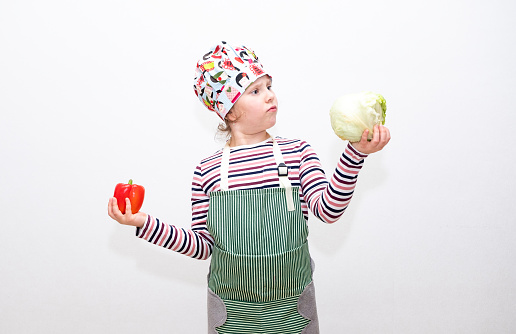 An 8-year-old Caucasian girl holds vegetables, red peppers and salad in her hands and thinks about what to choose, portrait on a light background.