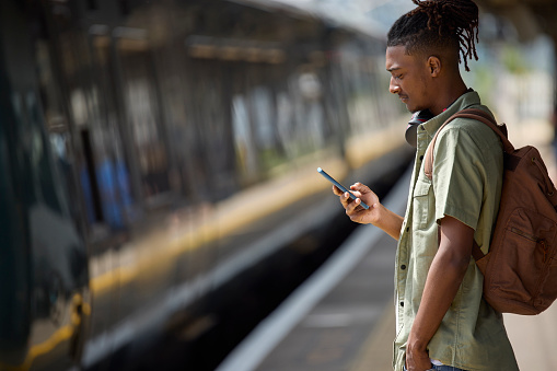 Young Man Commuting To Work On Train Standing  On Platform Looking At Mobile Phone As Train Arrives