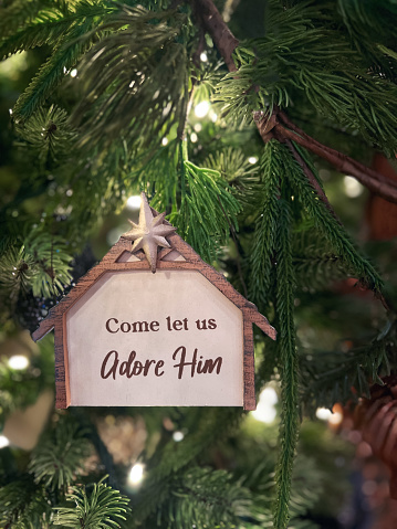 Religious ornament hanging from a brightly lit Christmas tree. The house or manger shaped ornament has the following words on it come let us adore him.