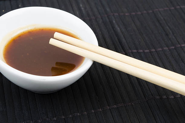 Hoisin &amp; Chopsticks Hoisin & Chopsticks - Close up of chopsticks resting on a small white bowl of asian dipping sauce on a black mat. hoisin sauce stock pictures, royalty-free photos & images